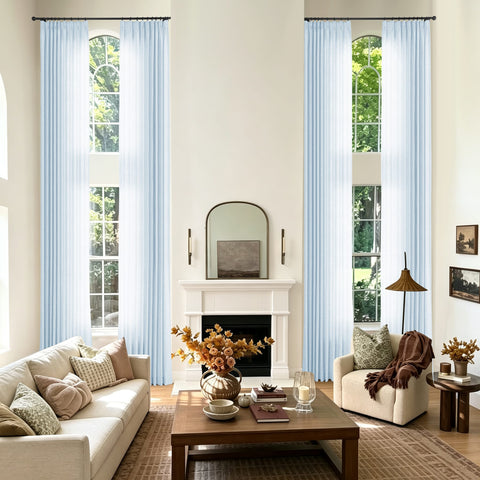 A styled living room with two tall windows dressed in light blue sheer curtains featuring a cream sofa and a chair with a throw blanket